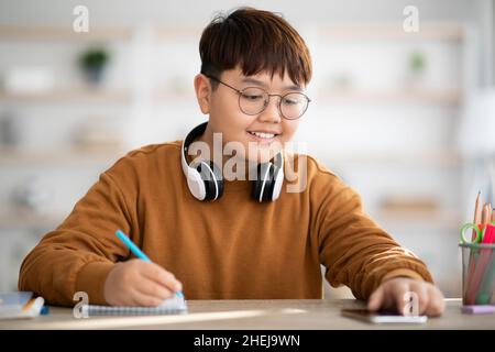 Smiling japanese kid with headset sitting at table, using smartphone Stock Photo