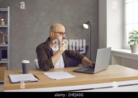 Serious businessman sitting at office desk, looking at laptop screen and thinking Stock Photo
