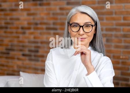 Close-up portrait of contemporary senior woman wearing stylish eyewear looking at the camera and smiles. Elderly female resting at the office, spending calm break Stock Photo