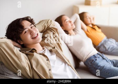 Happy youngsters japanese girl, millennial mother relax, father with hand behind head look at camera on couch Stock Photo