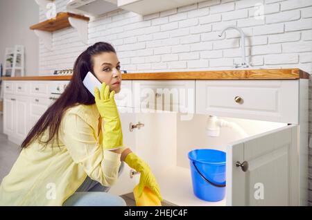Young woman calls the plumber as she has water leaking from her kitchen sink pipe Stock Photo