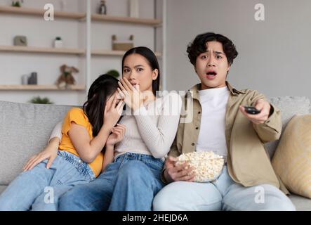 Frightened shocked asian millennial wife, husband and adolescent daughter watching scary movie and eating popcorn Stock Photo