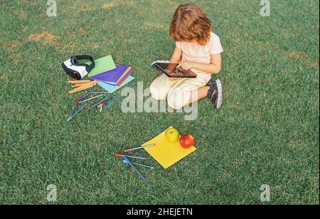 Child study exam outside. Funny kid little student boy with tablet, Sit on lawn in park, studying at School backyard. Stock Photo