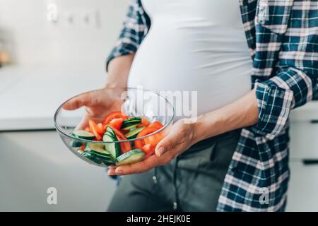 Cropped image pregnant woman hold bowl with vegetable salad of cucumbers and tomatoes in kitchen. Stock Photo