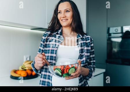 Portrait of young smiling pregnant woman mix vegetable salad with cucumbers and tomatoes in transparent bowl in kitchen. Stock Photo