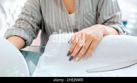 Female hands with artificial acrylic nails during manicure procedure. Colored nail polish on manicure in beauty salon. Client dries nails in lamp in Stock Photo