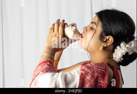A beautiful Indian woman in red saree blowing conch shell on white background Stock Photo