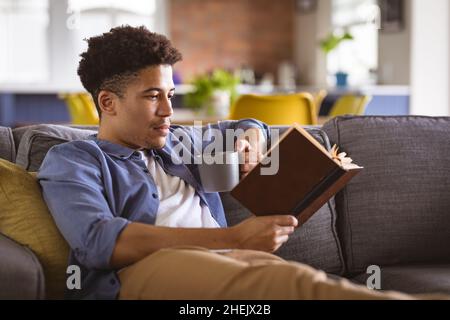 Young biracial handsome man holding coffee mug reading book while relaxing on sofa at home Stock Photo