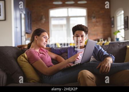 Young woman pointing at laptop while sitting with boyfriend on sofa in living room Stock Photo