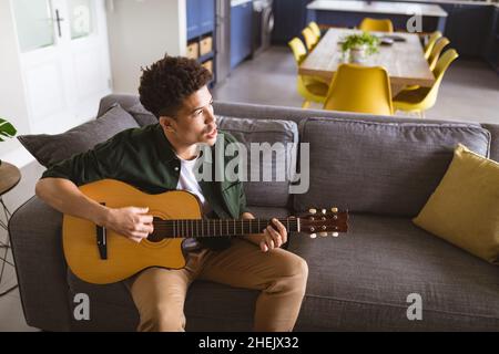 Young handsome biracial man practicing acoustic guitar and singing on sofa in living room Stock Photo