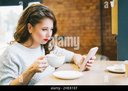 Concentrated young brunette female with cup of coffee in hands messaging on phone while sitting at table in comfortable cafe during lunch time break. Stock Photo
