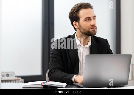 Pensive young businessman in formal wear sitting at desk in office, using laptop computer, watch away thoughtfully, looking for business tasks solving, male office employee in suit on the workplace Stock Photo
