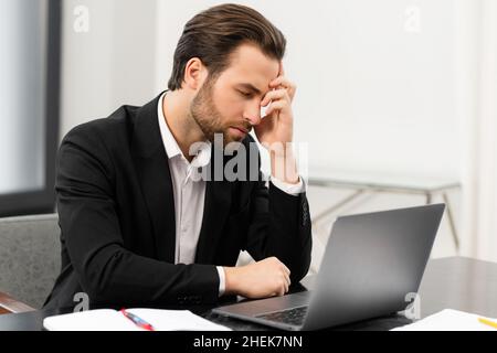 Frustrated young businessman in formal suit made mistake and worried about it, tired male office employee suffering from strong headache, sitting at the desk in office with eyes closed Stock Photo