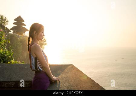 Bali - Indonesia - 10.21.2015: Asian young female with braided dark hair watching the sea at sunset - golden hour at Uluwatu Temple - side view Stock Photo