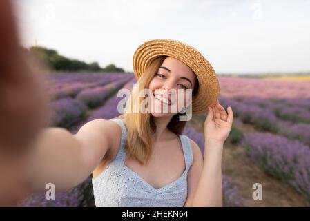 outdoors romantic portrait of young happy and attractive woman in white summer dress taking selfie with mobile phone on beautiful lavender flowers Stock Photo