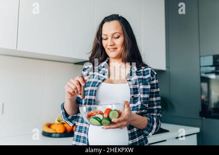 Portrait of young smiling pregnant woman mix vegetable salad with cucumbers and tomatoes in transparent bowl in kitchen. Stock Photo