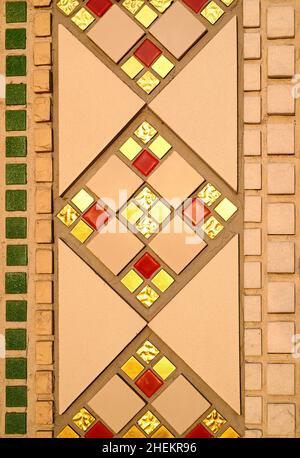 A fragment of a geometric repeating pattern from a ceramic mosaic on the wall of a Catholic church. Gold, red, beige and green mosaic tile. Stock Photo