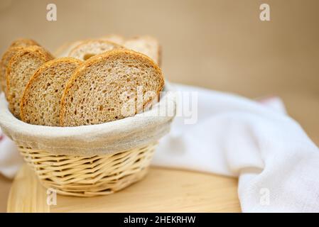 Fresh cereal bread in a wicker basket. The bread is sliced. Beige background, wooden board, linen cloth in a basket, white tea towel on the table. Stock Photo
