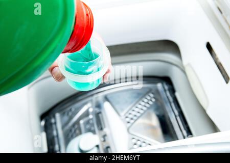 Liquid detergent from a green bottle is poured into the washing machine. Preparation for washing, measuring cup for washing powder. Stock Photo