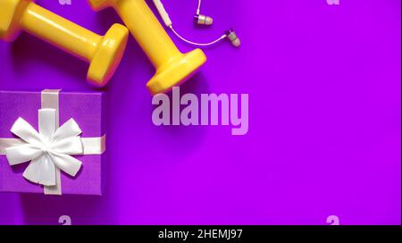 Christmas sports composition with dumbbells, small ear headphones, purple gift box with white bow on purple background. Stock Photo