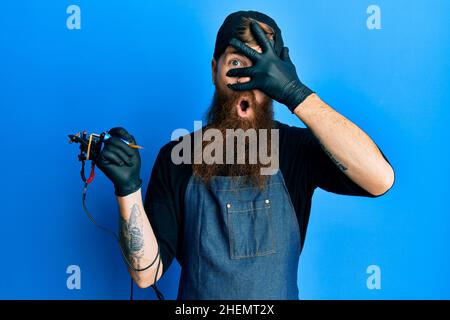 Redhead man with long beard tattoo artist wearing professional uniform and gloves peeking in shock covering face and eyes with hand, looking through f Stock Photo