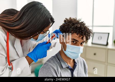 Young latin doctor woman auscultating the ear of man using otoscope at examination room. Stock Photo