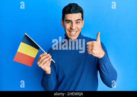 Handsome hispanic man holding belgium flag smiling happy and positive, thumb up doing excellent and approval sign Stock Photo