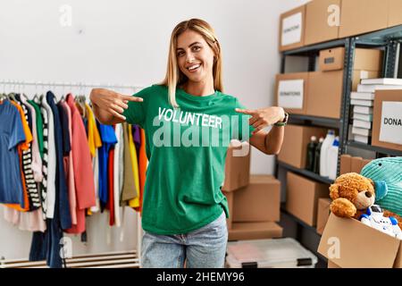 Young caucasian girl smiling happy pointing with fingers at volunteer uniform at charity center. Stock Photo