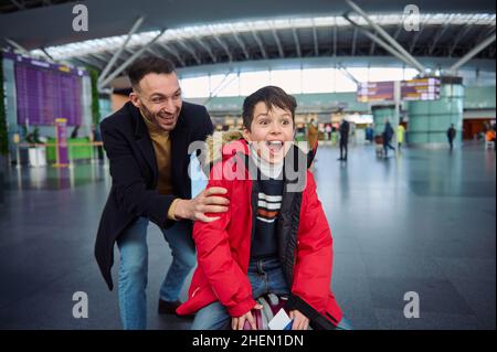 Cheerful happy young father rides his son on a suitcase in the departure hall of the international airport. Dad and son having fun together while wait Stock Photo