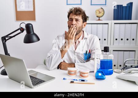 Young hispanic dentist man working at medical clinic bored yawning tired covering mouth with hand. restless and sleepiness. Stock Photo