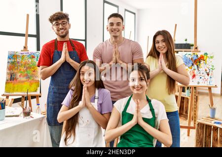 Group of five hispanic artists at art studio praying with hands together asking for forgiveness smiling confident. Stock Photo