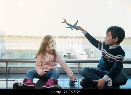 Handsome kids, boy and little girl playing with toys planes sitting on the floor by the panoramic windows overlooking the runways and planes in the de Stock Photo