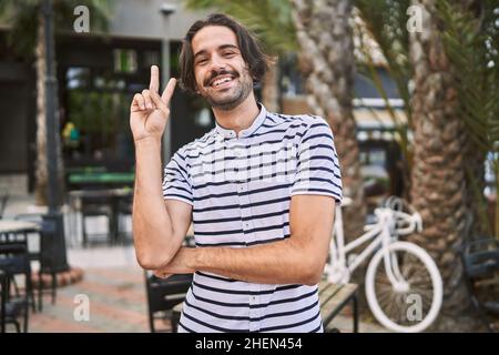 Young hispanic man with beard outdoors at the city smiling with happy face winking at the camera doing victory sign. number two. Stock Photo