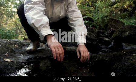 Tourist man washing his face in a mountain river in dense forest in a sunny spring day. Man collects fresh water from a spring in folded hands, drinks Stock Photo