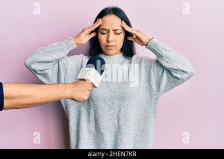 Young hispanic girl being interviewed by reporter holding microphone with hand on head, headache because stress. suffering migraine. Stock Photo