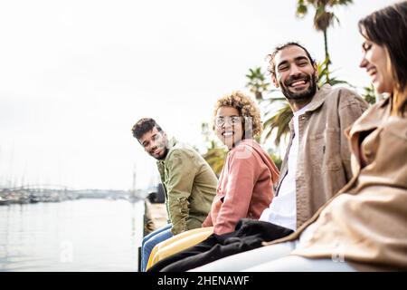 Young group of diverse adult friends laughing while sitting in front of the sea Stock Photo
