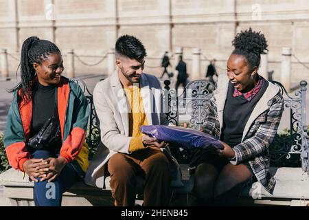 Three friends exchanging presents while sitting on a bench outdoors Stock Photo