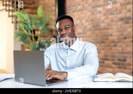 Positive confident black man using laptop in modern office. Portrait successful businessman, freelancer, manager, in formal shirt, working and typing on laptop, looking at camera, smiling friendly Stock Photo
