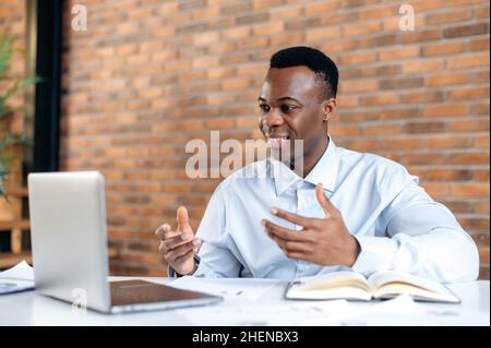 African american positive successful business man in shirt, ceo company, manager, broker, uses laptop for remote communication with client, sitting in modern office, gesturing with his hands, smiling Stock Photo
