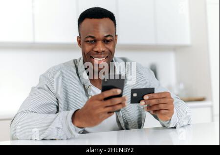 Positive african american young man sitting on kitchen at home, dressed in casual wear, using smartphone and credit card for online shopping, is entering data for payment, online transaction, smiles Stock Photo