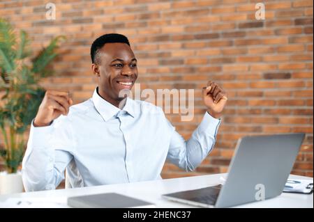 Success, win, luck. African american excited man, broker, agent, manager, working on laptop in modern office, amazed looks at screen, enjoying good deal, big profit, great news, gesturing with fists Stock Photo