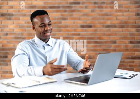 Remote conversation. African american successful business man, ceo company, sales manager, uses laptop for distant communication with client, sits in modern office, gesturing with his hands, smiling Stock Photo