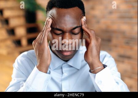 Close-up of an exhausted african american man experiencing headache, migraine, overwork from working at the computer, sad guy is sick, needs treatment and rest, closed his eyes, massages temples Stock Photo