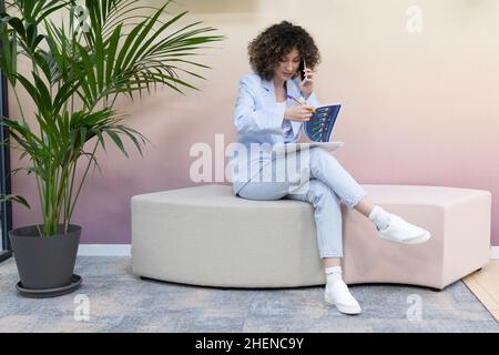 Happy female office worker sitting in a couch talking on the phone and taking notes. Stock Photo