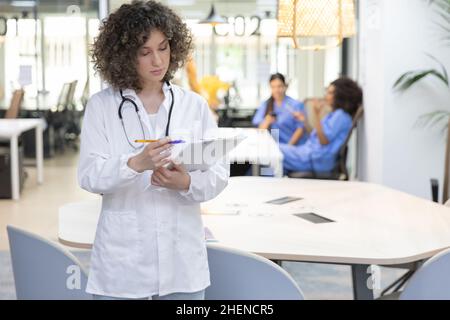 Concentrated female doctor with curly hair working in an office. In the background other medical workers are resting. Stock Photo