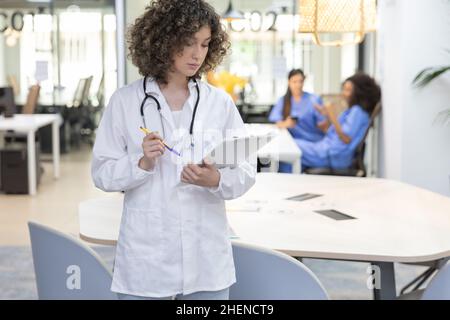 Concentrated female doctor with curly hair working in an office. In the background other medical workers are resting. Stock Photo