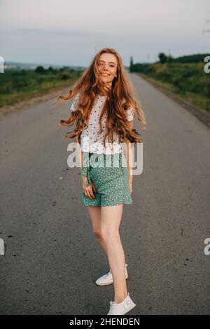 Redhead woman walking down on rural road near wheat field smiling at camera. Beautiful girl. Wheat field. Beautiful summer landscape. Stock Photo
