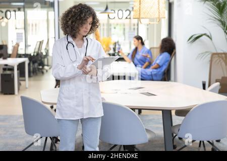 Concentrated female doctor with curly hair working in an office. In the background other medical workers are resting. Stock Photo