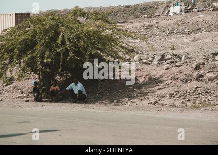 Djibouti, Djibouti - May 21, 2021: A Djiboutian man and his two sons sitting on the stone and waiting the bus under a tree's shade in Djibouti. Editor Stock Photo