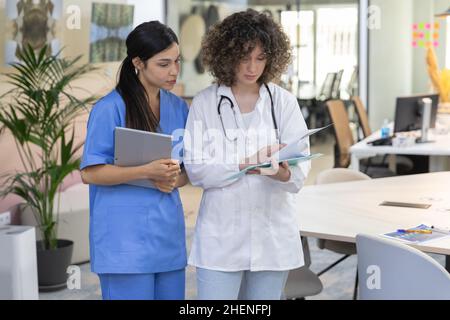 Concentrated female doctor with curly hair talking with a nurse and looking at paperwork. Stock Photo